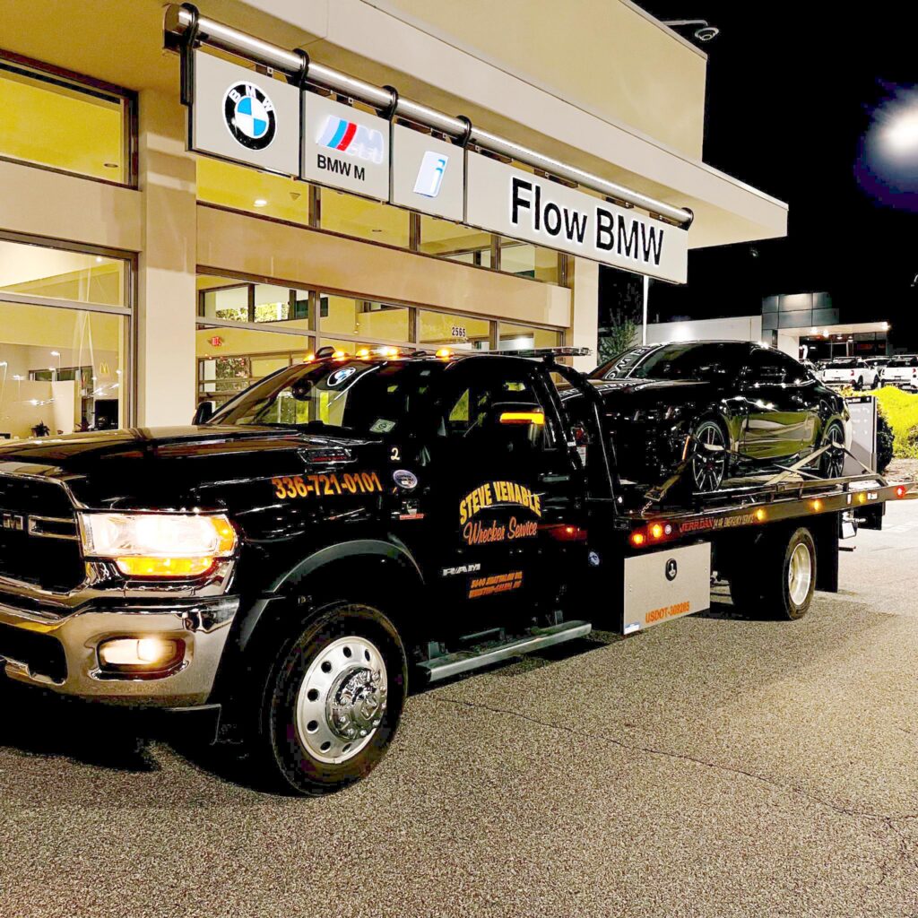 Nighttime view of Flow BMW dealership with BMW logos and a car carrier trailer in front of the building, several black trucks visible below the sign.